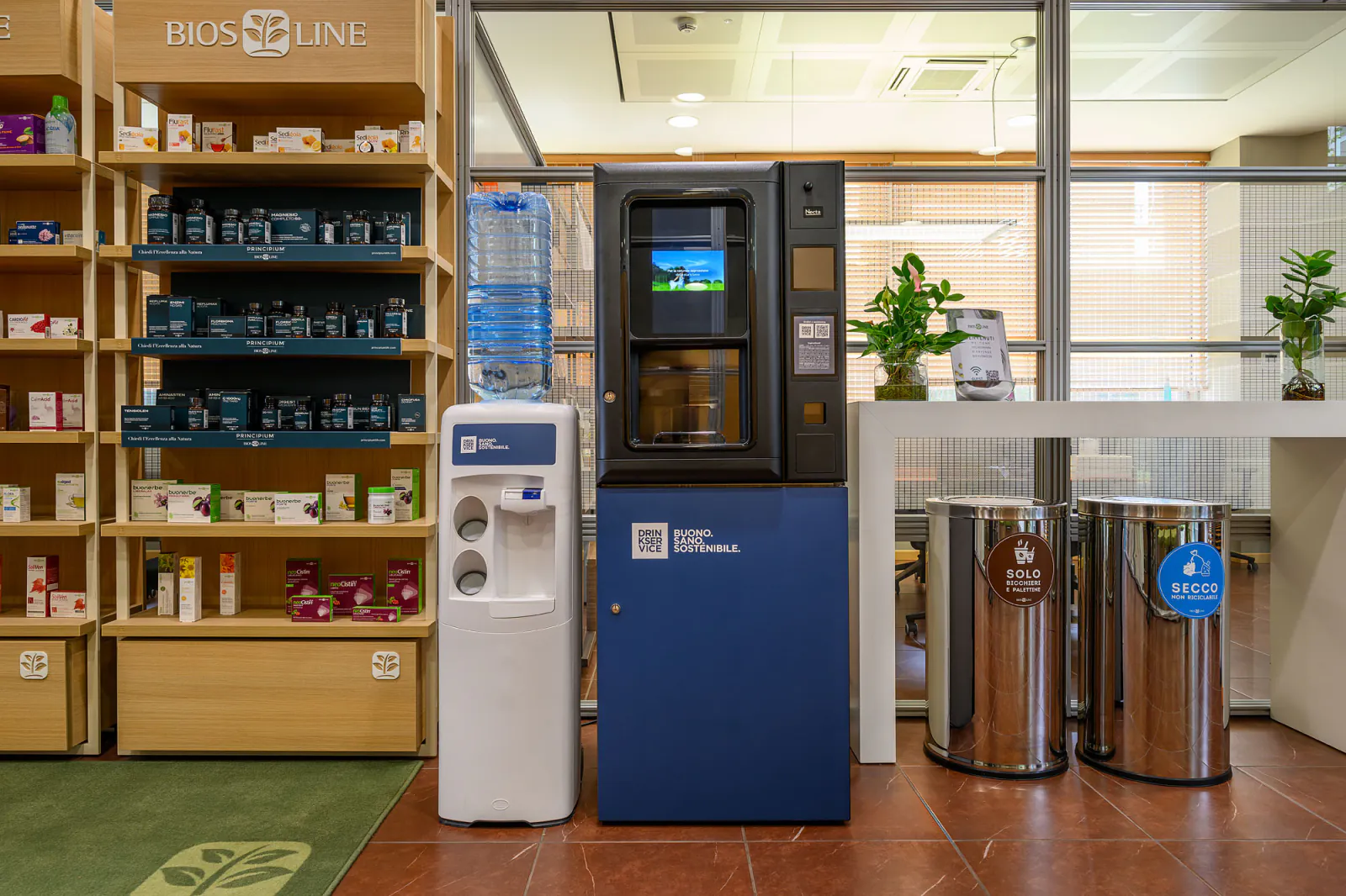 Distributore e dispenser a boccione affiancati sotto l'insegna aziendale, fra gli scaffali in legno del punto vendita e due cestini per la differenziata.