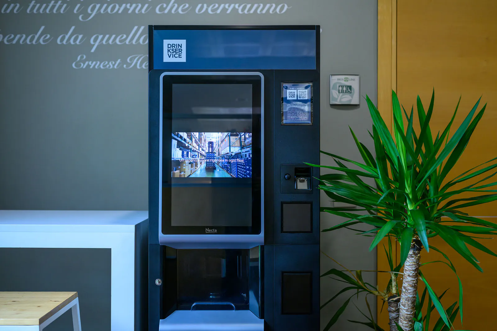 Veduta d'angolo dell'area pausa, con il tavolo bianco in primo piano e il distributore acceso di fianco alla porta.