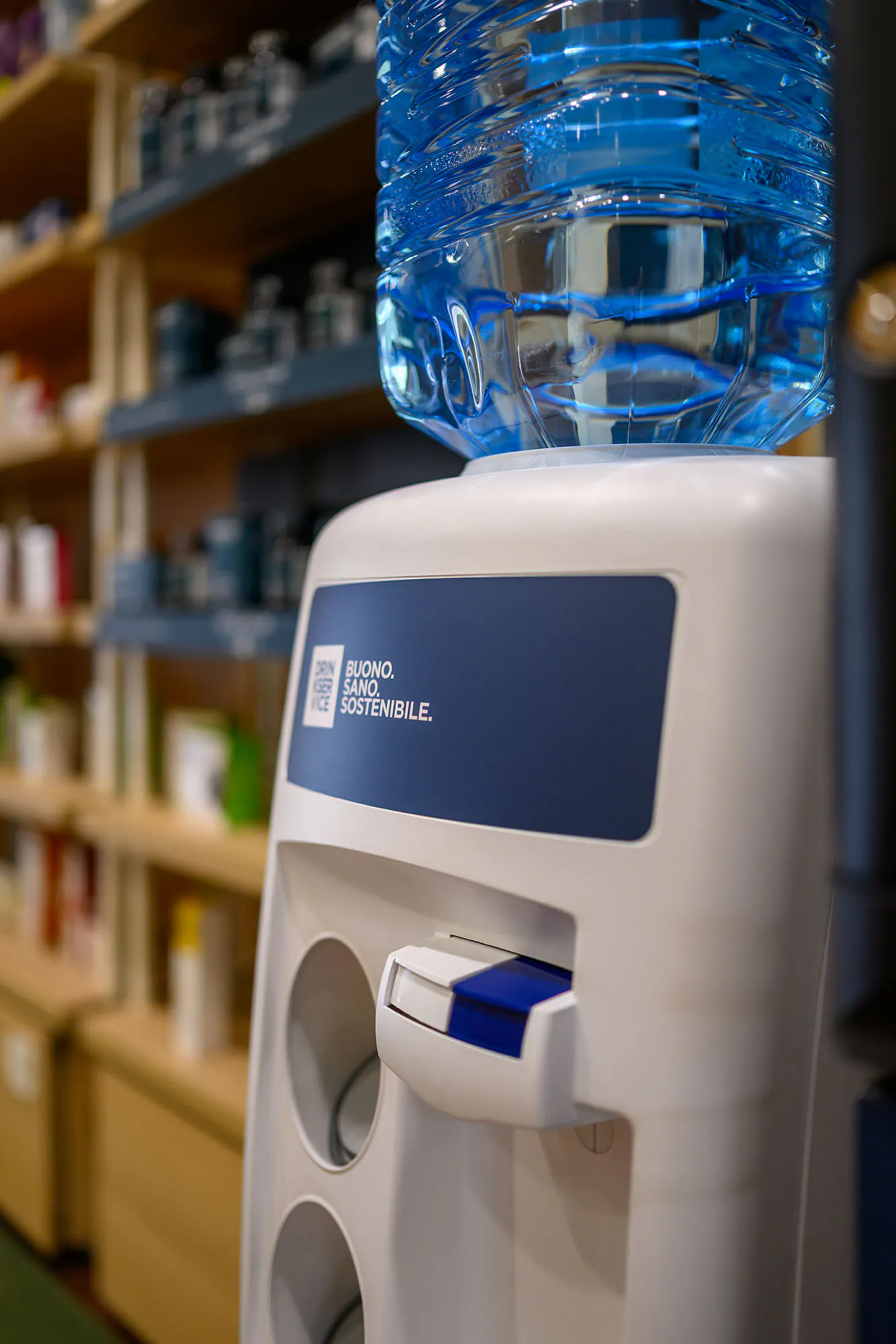 Dispenser con boccione d’acqua accanto al distributore, davanti agli scaffali in legno del punto vendita.