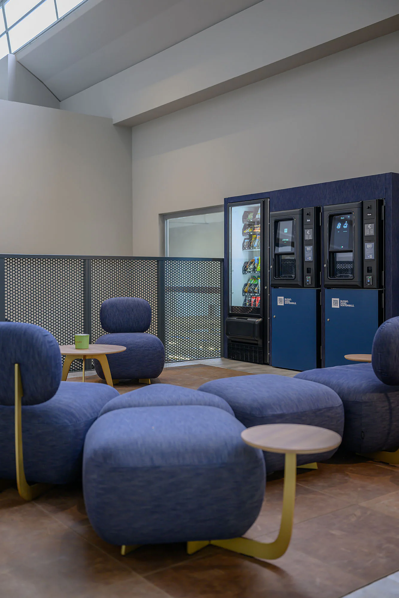 Area break su un ballatoio con lucernari: pouf e poltrone in denim blu su un tappeto, tavolini tondi con gambe dorate e i distributori sul fondo.