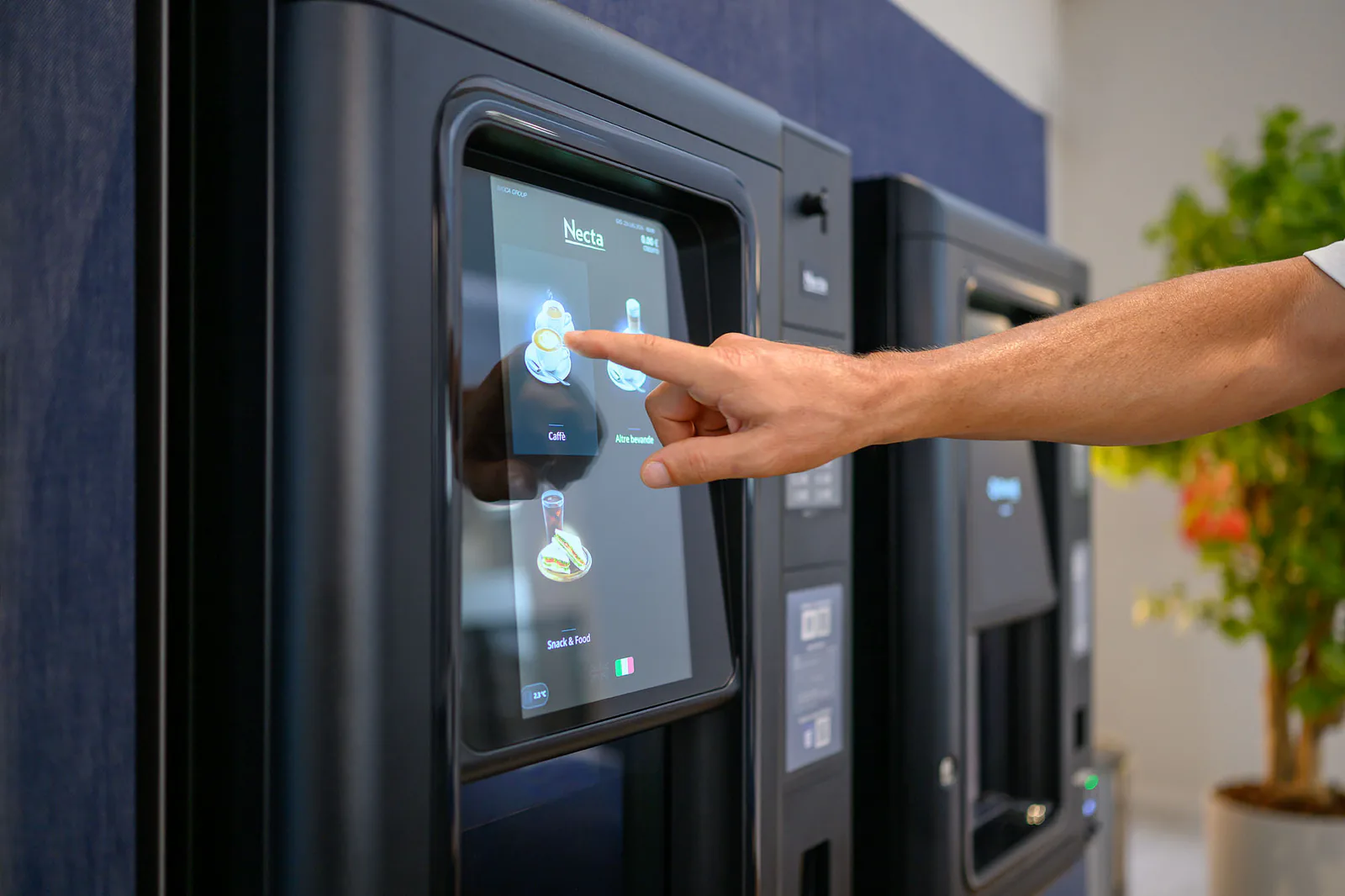 Un braccio si allunga a toccare il touchscreen del distributore, con un agrume in vaso sullo sfondo.