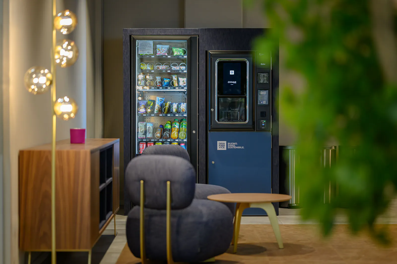 Angolo pausa di sera: lampade a sospensione dorate, mobile in legno, poltrona in denim e i due distributori accesi.