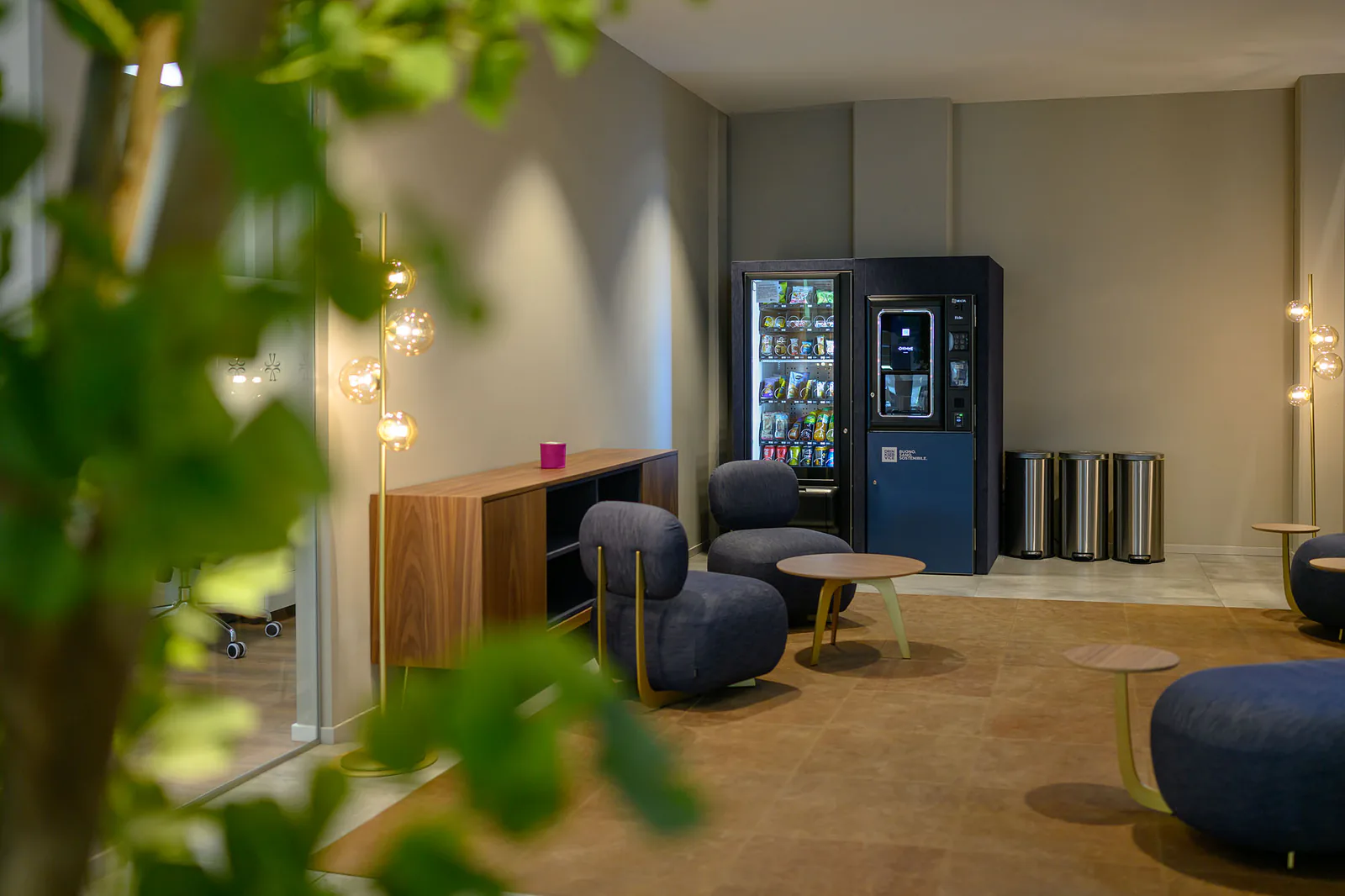 La lounge ripresa attraverso il fogliame di un albero da interno, con il filare di lampade e i distributori in fondo.