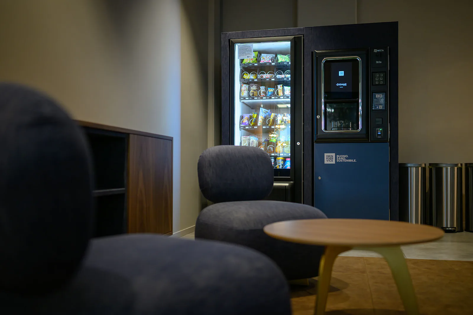 Poltrona in denim e tavolino tondo in primo piano, con il distributore di snack illuminato alle spalle.