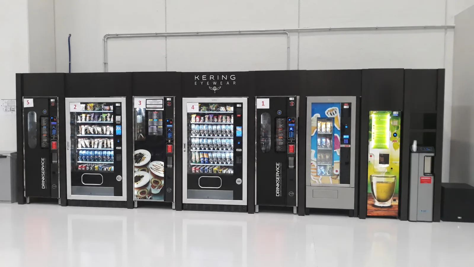Batteria di otto distributori automatici affiancati e numerati da 1 a 5, inseriti in un mobile nero su misura sotto l'insegna aziendale, in un ambiente industriale con pavimento lucido.