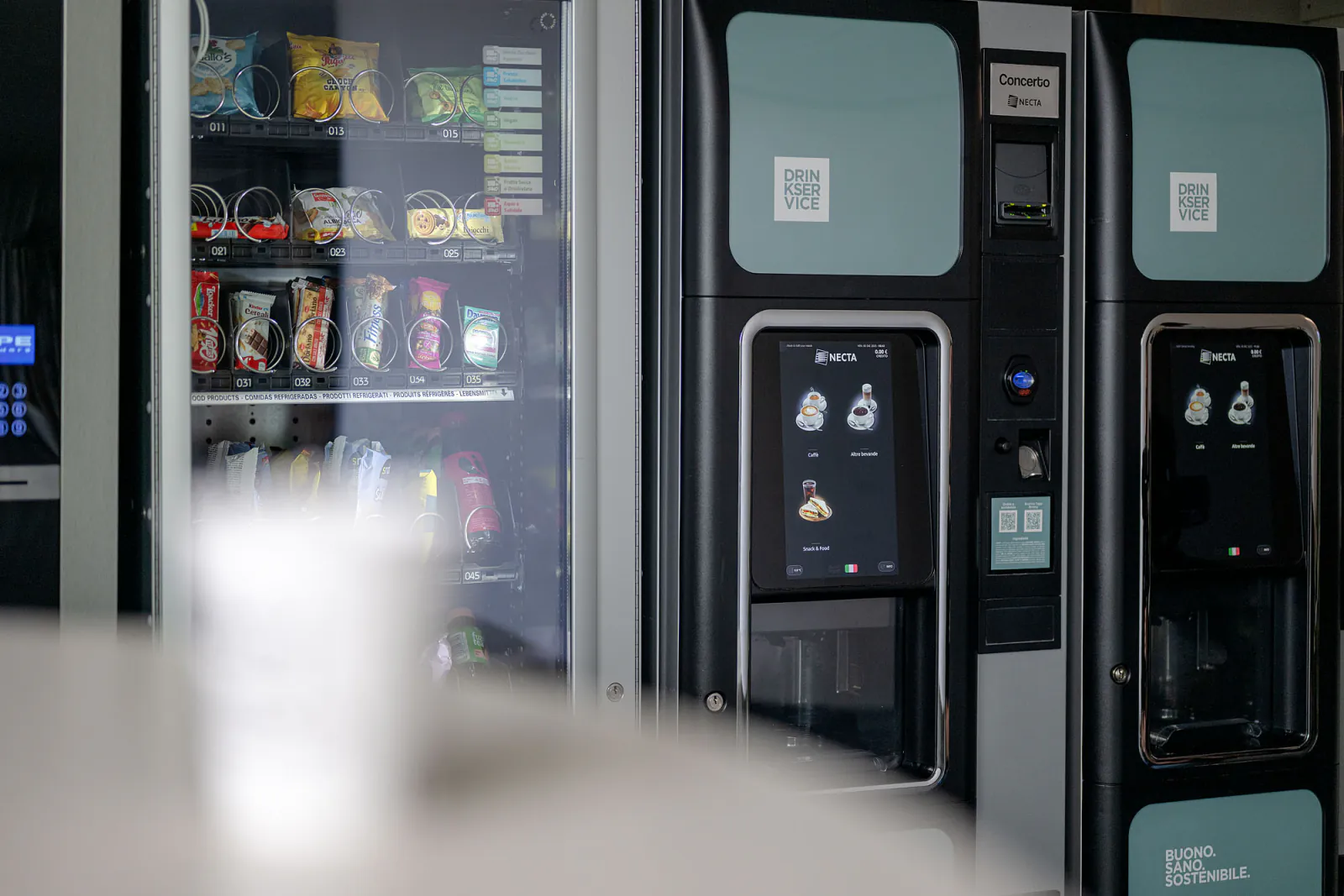 Distributori di bevande, snack e acqua nella hall aziendale, con piante e arredi dell’area ristoro.