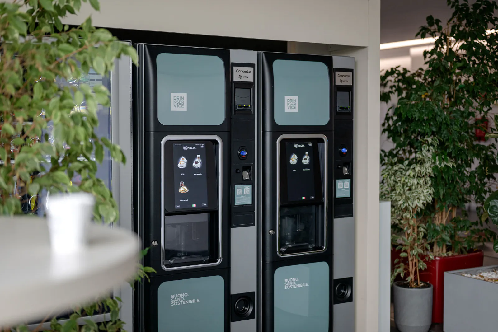 Distributori di bevande, snack e acqua nella hall aziendale, con piante e arredi dell’area ristoro.