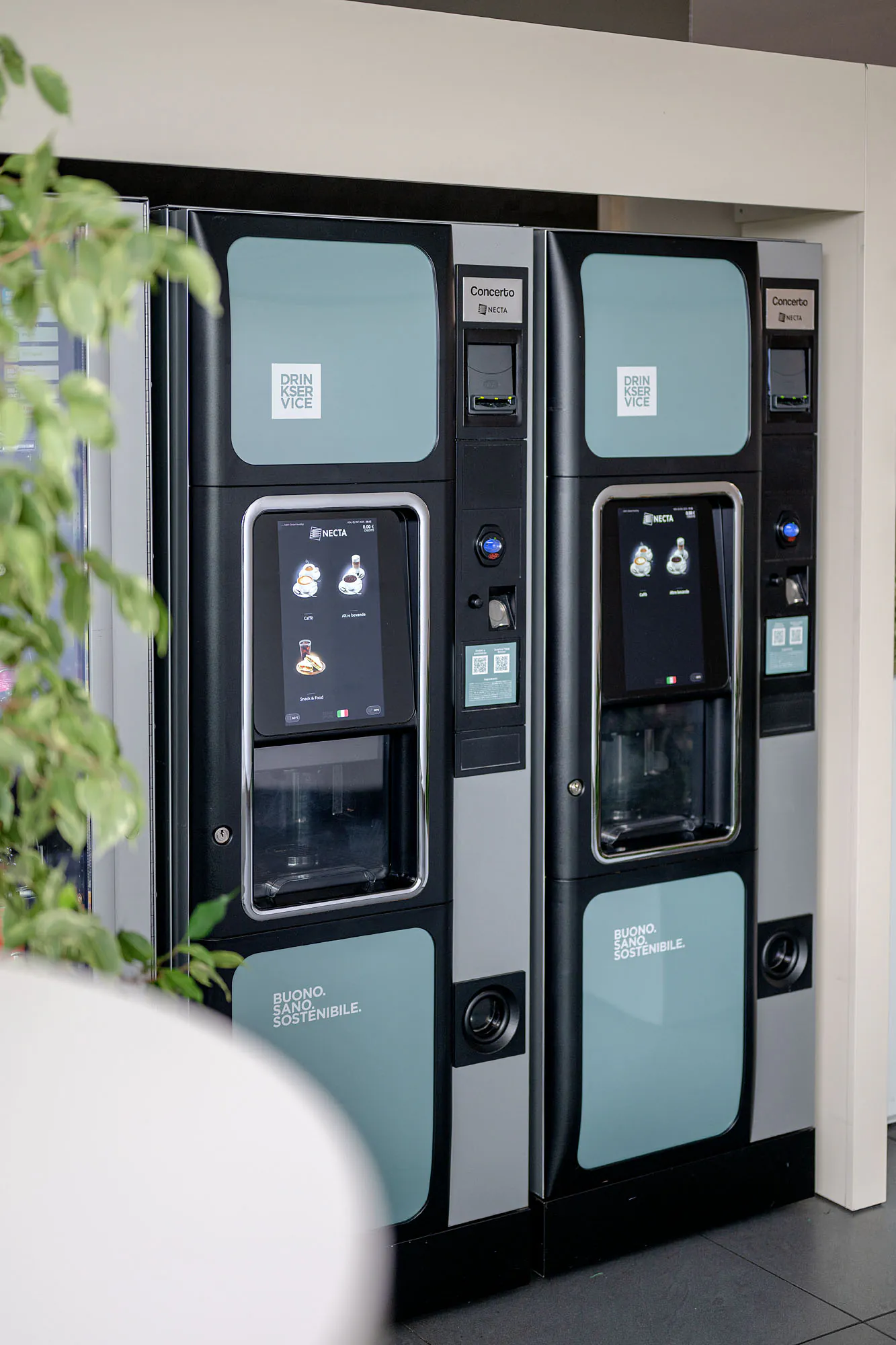 Distributori di bevande, snack e acqua nella hall aziendale, con piante e arredi dell’area ristoro.