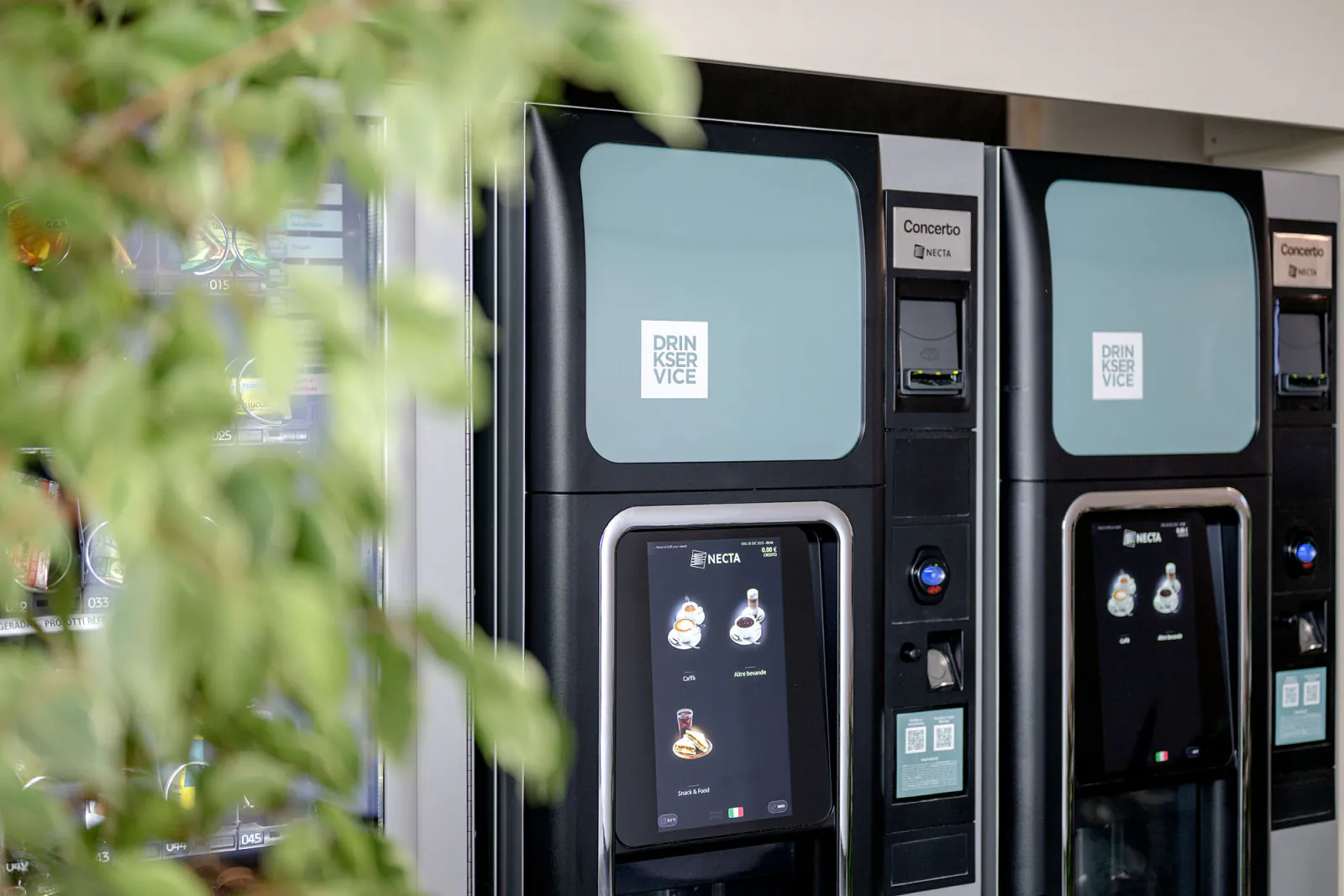 Distributori di bevande, snack e acqua nella hall aziendale, con piante e arredi dell’area ristoro.