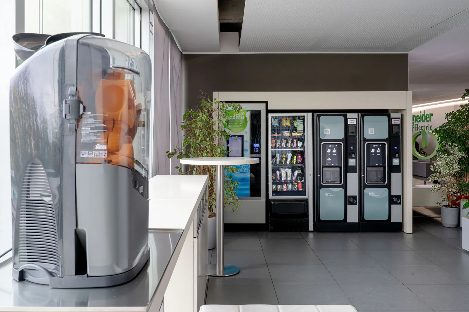 Distributori di bevande, snack e acqua nella hall aziendale, con piante e arredi dell’area ristoro.