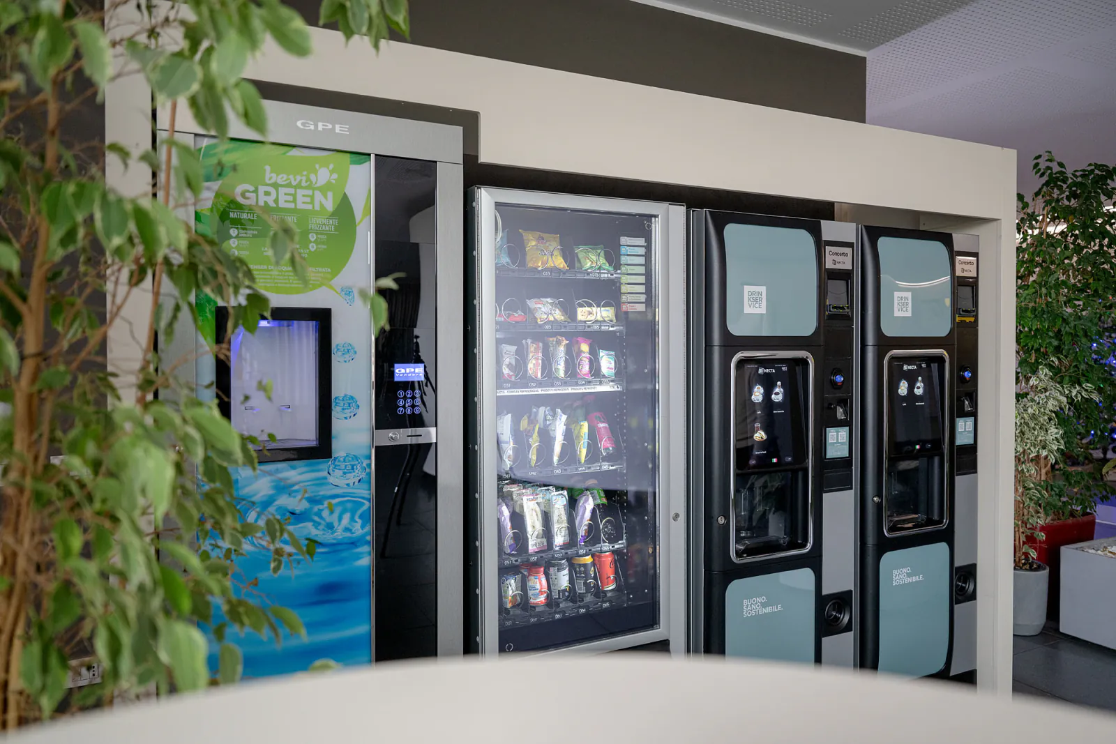 Distributori di bevande, snack e acqua nella hall aziendale, con piante e arredi dell’area ristoro.