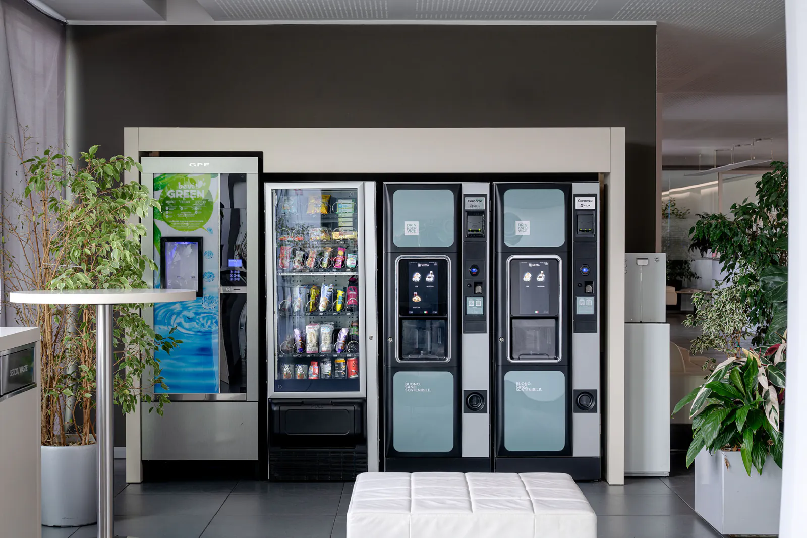 Distributori di bevande, snack e acqua nella hall aziendale, con piante e arredi dell’area ristoro.